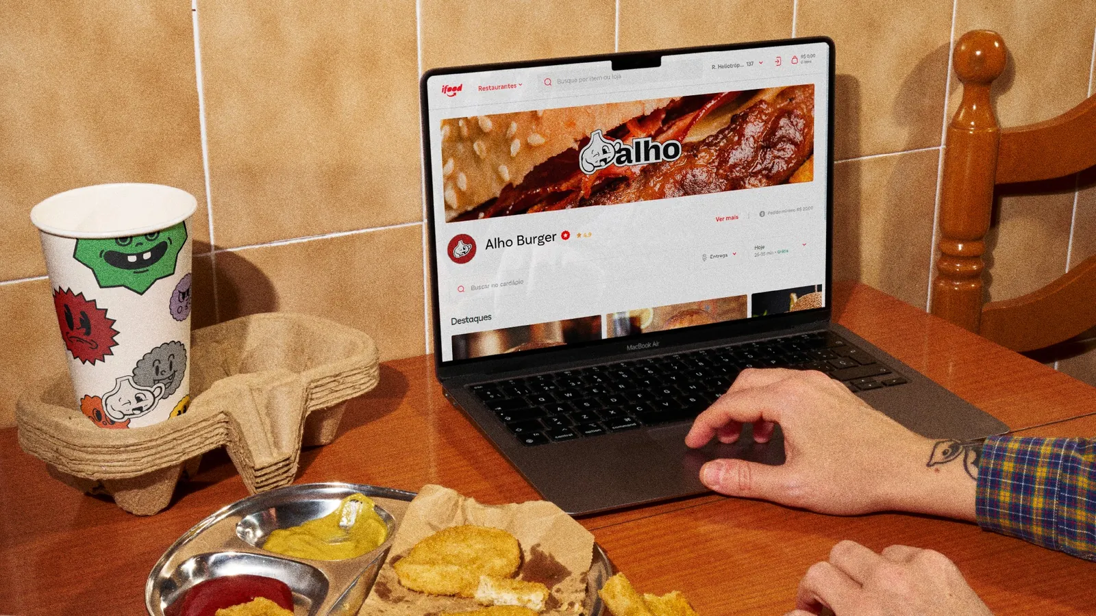 Notebook com a página da Alho Burger no iFood, copo com o mascote de alho e lanche na mesa